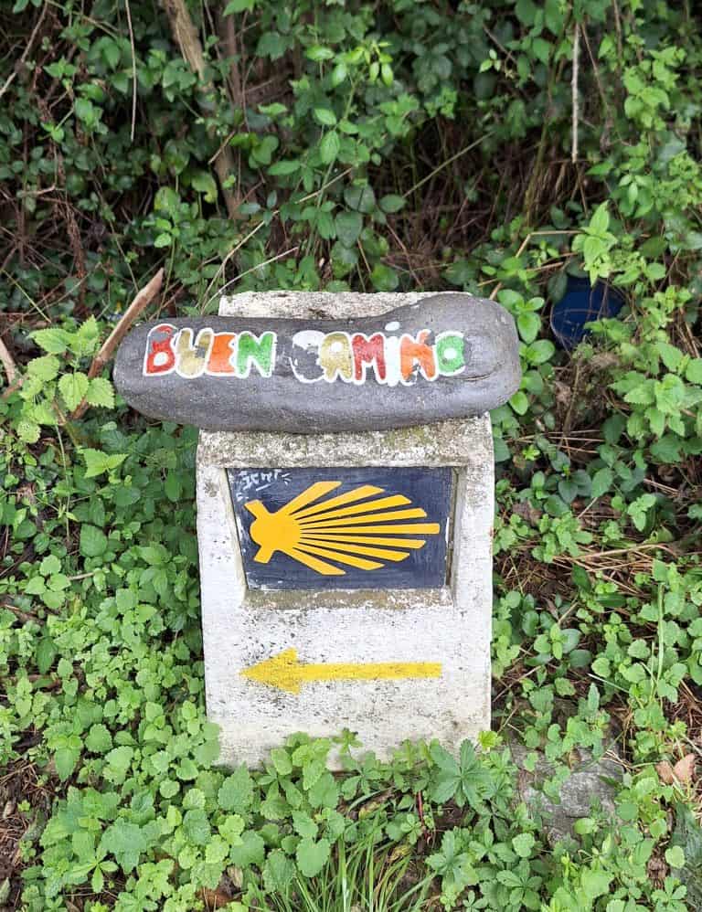 “Buen Camino” stone resting on a Camino de Santiago marker with scallop shell and yellow arrow, surrounded by lush grass.