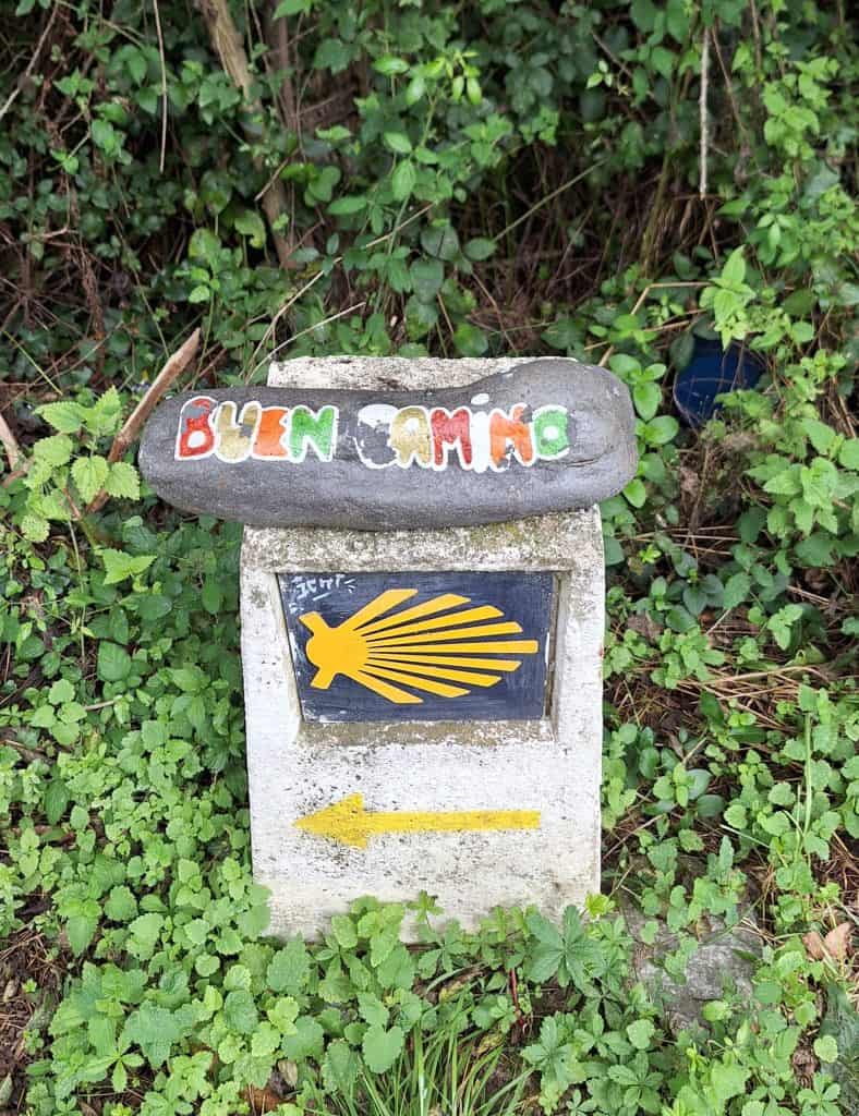“Buen Camino” stone resting on a Camino de Santiago marker with scallop shell and yellow arrow, surrounded by lush grass.